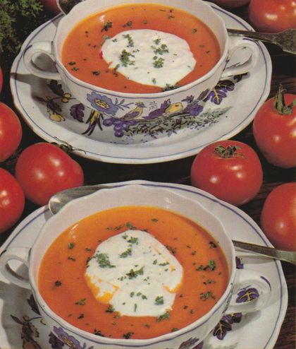 Zwei Suppentassen mit blumenverziertem Porzellan enthalten orangefarbene Tomatensuppe, garniert mit einem Klecks Sahne und frischen Kräutern. Rundherum sind frische Tomaten, ein silberner Löffel und eine Serviette auf dunklem Untergrund arrangiert.