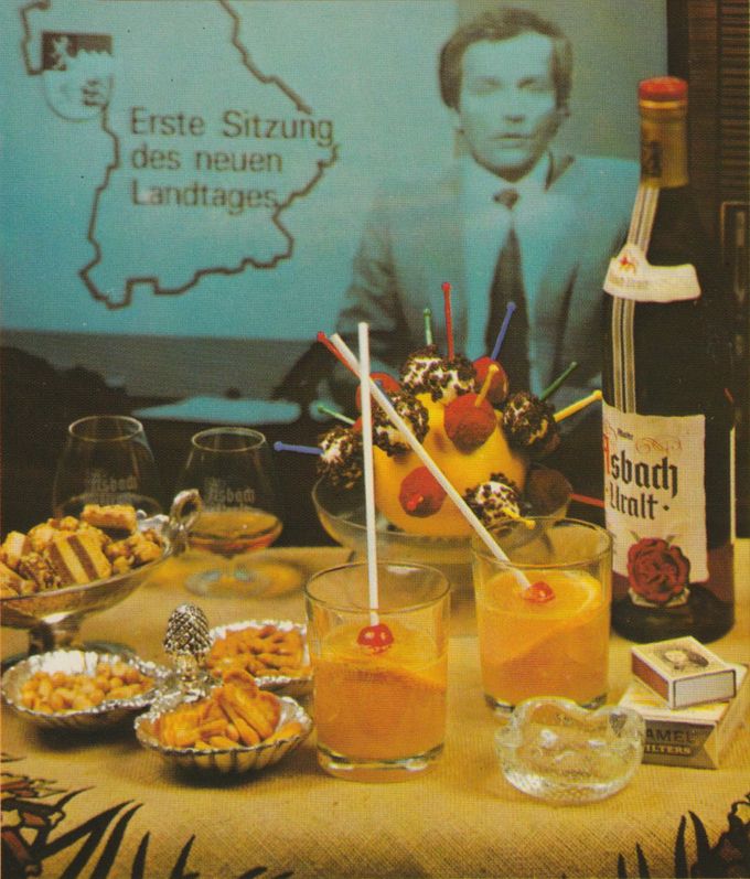 Ein festlich gedeckter Tisch mit Cocktails in Gläsern mit bunten Trinkhalmen, kleinen Gebäcktörtchen, einer Flasche Asbach-Uralt und einem dekorativen Arrangement mit bunten Spießen im Hintergrund. Im Hintergrund ist ein Fernsehbild mit der Aufschrift „Erste Sitzung des neuen Landtages“ zu sehen.