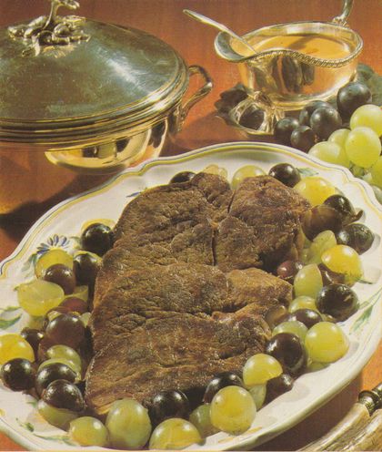 Vier gebratene Hirschsteaks auf einem ovalen Teller mit gelb-grünem Blumenmuster, umgeben von halbierten grünen und dunklen Weintrauben. Im Hintergrund eine goldene Servierschüssel mit Deckel und eine Sauciere mit Weintrauben-Dekoration.