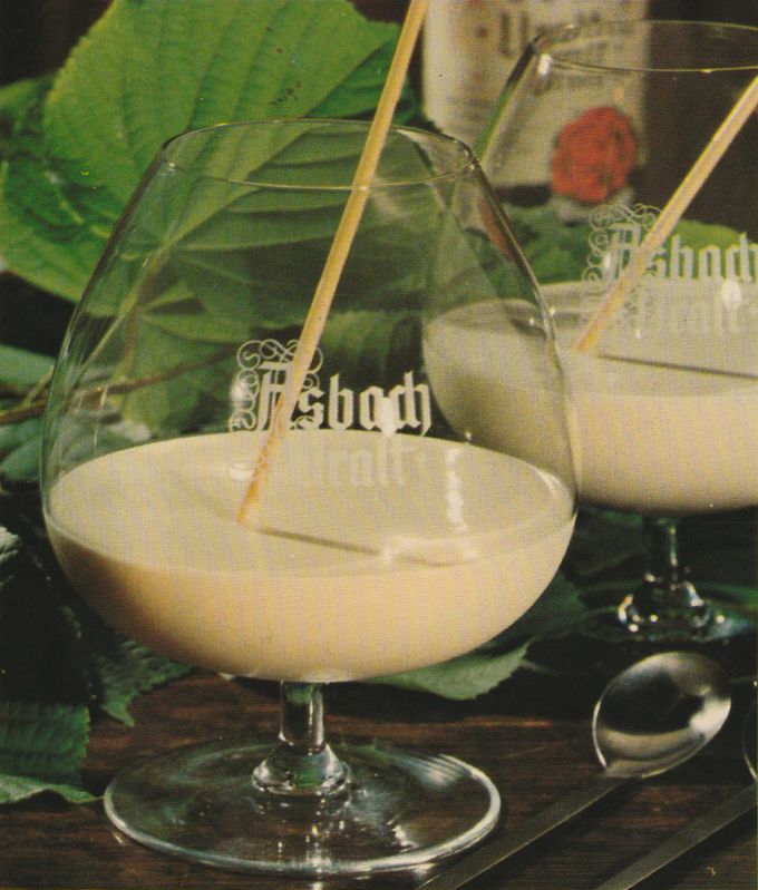 Zwei Cognac-Schwenker mit cremigem Eisgetränk, in das mit einem Strohhalm eingeschenkt wird. Im Hintergrund dekorative Blätter und eine Flasche Asbach Uralt. Neben den Gläsern liegt ein Löffel.