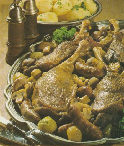Gebratene Gänsekeulen auf einem Bett aus Champignons, Schalotten und Maronen, garniert mit kleinen Bratwürstchen. Im Hintergrund eine Schüssel mit Salzkartoffeln, bestreut mit Petersilie, sowie Salz– und Pfeffermühle. Die Speisen sind auf einer ovalen Servierplatte angerichtet.