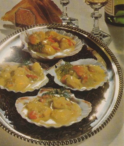 Vier geöffnete Jakobsmuscheln in ihren Schalen liegen auf einem silbernen Tablett, serviert in einer cremigen gelben Sauce mit Garnelen und frischem Dill garniert. Im Hintergrund sind Kristallgläser und getoastetes Brot zu sehen.