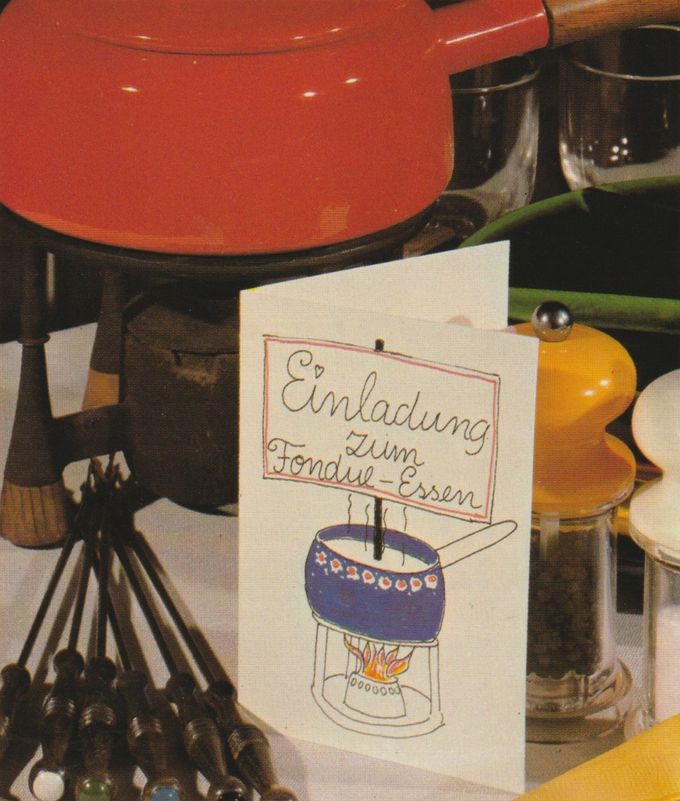 Eine handgeschriebene Einladungskarte zum Fondue-Essen lehnt vor einem orangefarbenen Fondue-Set mit schwarzem Rechaud. Daneben liegen Fonduegabeln, ein Pfeffer- und ein Salzstreuer.