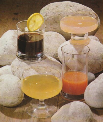 Vier verschiedene Cocktails in unterschiedlichen Gläsern, arrangiert auf hellen Steinen. Links hinten ein dunkler Drink mit Zitronenscheibe am Glasrand, daneben ein cremefarbener Cocktail in einer Sektschale. Vorne links ein Drink in einer bauchigen Cocktailschale, rechts daneben ein Glas mit einem Getränk in Orange– und Rottönen.