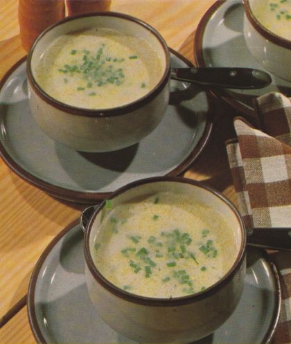 Zwei Suppentassen mit cremiger, heller Suppe, garniert mit gehackten Kräutern, stehen auf Untertassen neben einer karierten Serviette und sind auf einem hellen Holztisch arrangiert.