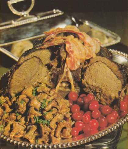 Gebratener Hirschrücken auf ovaler Servierplatte, belegt mit knusprigen Frühstücksspeck-Scheiben. Dazu arrangiert: Semmelknödel, gebratene Pfifferlinge mit Petersilie und rote Johannisbeeren als Dekoration. Im Hintergrund weitere Beilagen auf Tellern.