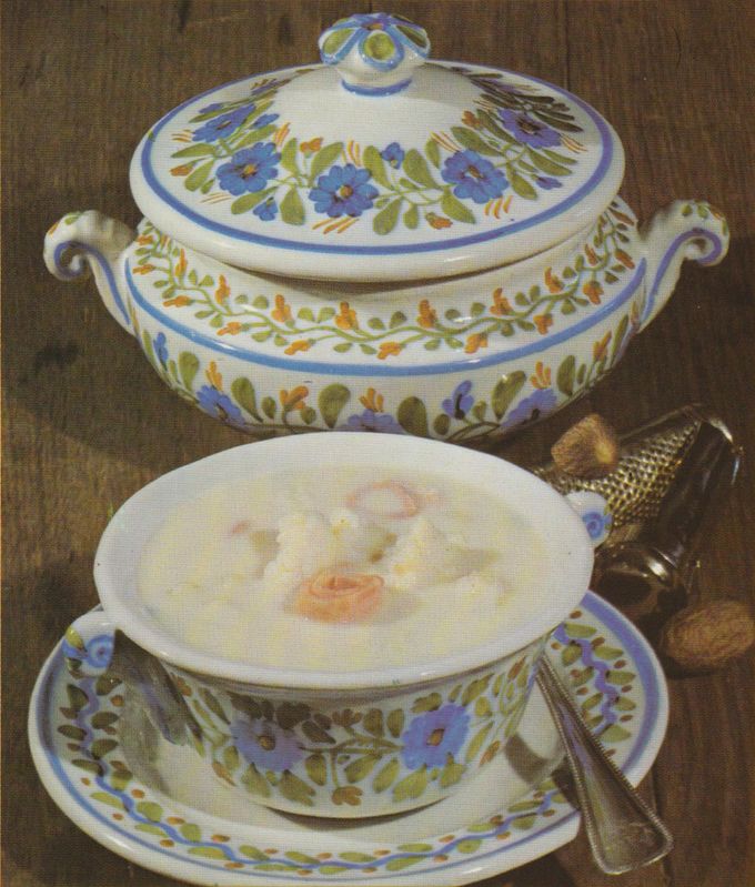 Eine cremige Suppe wird in einem Suppenteller mit passender Untertasse serviert, das Geschirr ist mit blauem Blumenmuster und grün-orangen Blättern verziert. Im Hintergrund steht eine große Suppenterrine mit Deckel im gleichen Dekor, rechts daneben liegt eine Muskatnussreibe.