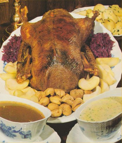 Gebratene Gans auf einer Servierplatte, umgeben von gedünsteten Apfelspalten, Maronen und Rotkohl. Im Hintergrund eine Schüssel mit Salzkartoffeln. Im Vordergrund zwei Saucieren mit heller und dunkler Sauce. Links hinten eine Kerze in einem Messingkerzenständer.