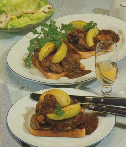 Zwei Tellergerichte mit gebratener Leber auf Toastbrot, garniert mit Apfelscheiben, gebratenen Zwiebeln und Petersilie, dazu Bratensoße. Im Hintergrund eine Glasschale mit grünem Blattsalat und ein Glas Weißwein.