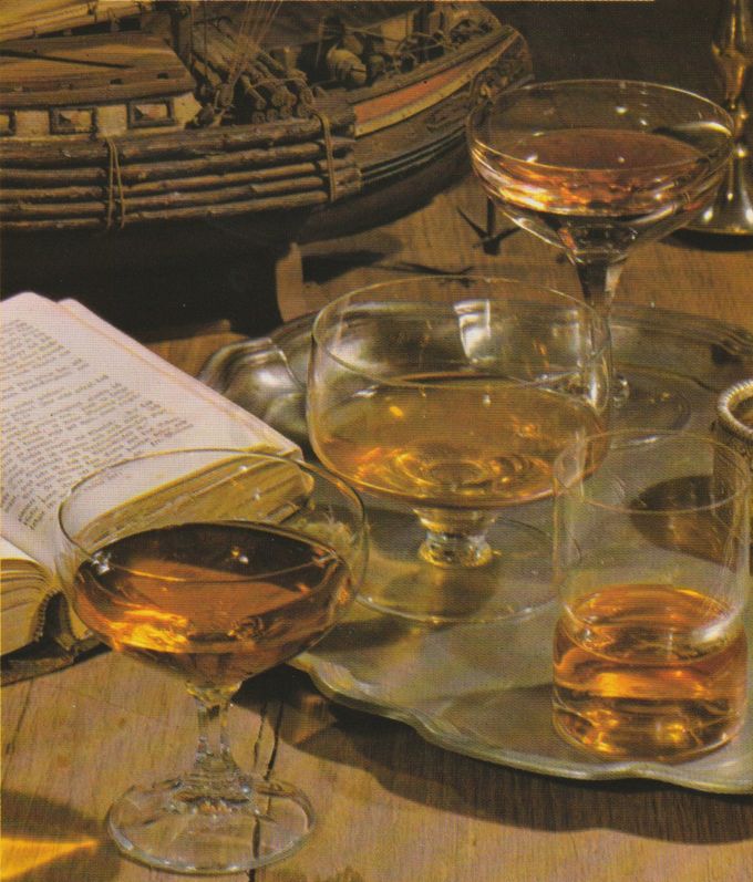 Verschiedene Gläser mit bernsteinfarbenen Spirituosen oder Likören stehen auf einem Holztablett neben aufgeschlagenen Büchern und geflochtenen Körben. Im Hintergrund ist rustikale Dekoration mit dem Modeel eines Schiffs aus Holz zu erkennen.