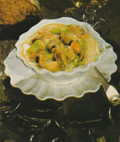 Eine cremige Suppe mit geschnittenen Lauchringen in einer weißen Porzellanterrine mit gewelltem Rand, serviert auf einem dunklen Holztablett. Im Hintergrund sind eine Tasse und eine Brot zu erkennen.