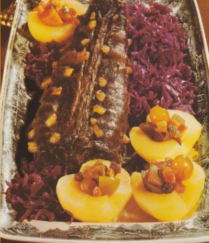 Gebratene Hasenrücken auf einer silbernen Servierplatte, garniert mit gelben Maiskörnern. Daneben gefüllte Apfelhälften mit kandierten Früchten und Rotkohl. Im Hintergrund eine weitere gefüllte Apfelhälfte auf dem Plattenrand.