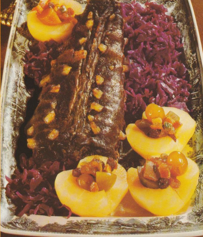 Gebratene Hasenrücken auf einer silbernen Servierplatte, garniert mit gelben Maiskörnern. Daneben gefüllte Apfelhälften mit kandierten Früchten und Rotkohl. Im Hintergrund eine weitere gefüllte Apfelhälfte auf dem Plattenrand.