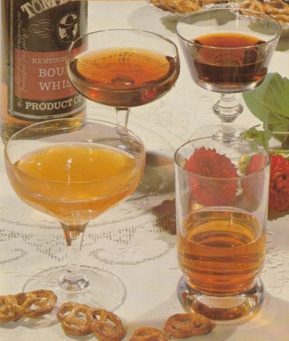 Vier verschiedene Cocktails in Coupe- und Tumbler-Gläsern mit bernsteinfarbenen bis dunklen Flüssigkeiten, neben einer Flasche Kentucky Bourbon Whiskey auf einer Spitzendecke. Im Vordergrund liegen Brezeln, rechts ist eine Nelke als Dekoration sichtbar.