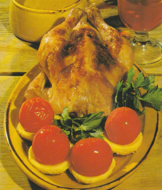 Knusprig gebratene Poularde auf ovalem Servierteller, garniert mit vier Tomaten auf Artischockenböden und frischer Petersilie. Im Hintergrund ein gefülltes Weinglas und eine Schüssel.