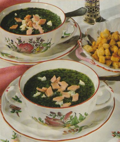 Zwei Suppentassen mit cremiger Spinatsuppe, garniert mit Schinkenwürfeln und Sahnetupfern, serviert auf Tellern mit Blumendekor. Daneben eine Schale mit goldbraun gebratenen Weißbrot–Croutons. Im Hintergrund Pfeffermühle und Serviette auf weißer Tischdecke.