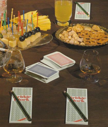 Ein gedeckter Spieltisch mit Käsewürfeln und Weintrauben auf Spießen, Salzgebäck auf einer Glasplatte, Schwenkern mit Cognac, Spielkarten und Bridge-Blöcken.