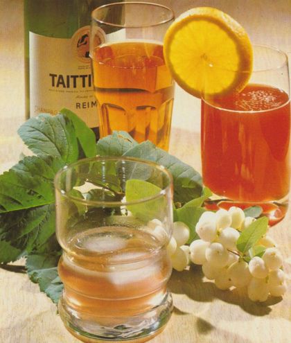 Drei verschiedene Getränke in Gläsern sind arrangiert mit frischen Weintrauben, grünen Weinblättern und einer halben Orange als Garnitur, im Hintergrund steht eine Flasche. Die Drinks zeigen sich in klarem Gelb, leuchtendem Orange und kräftigem Rot.