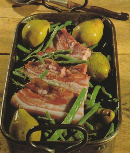 Ein gebratener Schweinebauch mit knuspriger Kruste liegt in einer dunklen Bratform, umgeben von grünen Bohnen und gelben Kartoffeln. Die Bratenschwarte ist mit ganzen Birnen und Bohnen garniert.