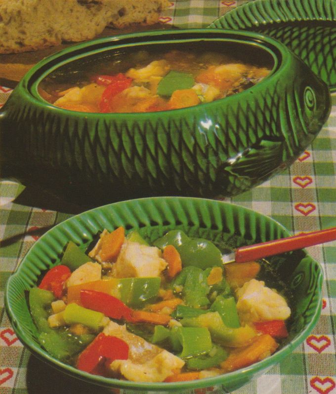 Gemüse–Fisch–Suppe in einer Schüssel und einer dekorativen Keramikschale in Fischform. Die Suppe enthält große Fischstücke, Paprikastreifen, Möhren, Lauch und Sellerie in klarer Brühe. Die Schüssel und Schale haben ein gerilltes Muster, karierte Tischdecke mit Herzmotiv im Hintergrund.