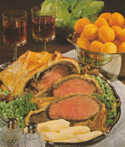 Festlich angerichtetes Rinderfilet im Blätterteig auf einer ovalen Servierplatte, umgeben von grünen Erbsen, Kartoffelkroketten. Im Hintergrund eine Schale mit Kroketten, zwei Gläser Rotwein, sowie grüner Salat.