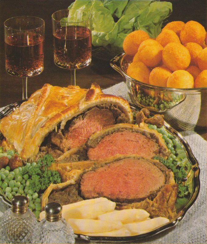 Festlich angerichtetes Rinderfilet im Blätterteig auf einer ovalen Servierplatte, umgeben von grünen Erbsen, Kartoffelkroketten. Im Hintergrund eine Schale mit Kroketten, zwei Gläser Rotwein, sowie grüner Salat.