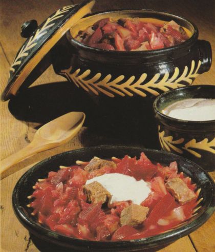 Ein Schmortopf aus dunkler Keramik mit gelbem Blattmuster enthält Fleischstücke mit Zwiebeln, daneben steht ein Teller mit Borschtsch mit Fleisch, Kohl und einem Klecks Sauerrahm. Rechts im Bild eine Schale mit weißer Creme oder Sauerrahm, links ein Holzlöffel auf einem gelben Geschirrtuch.