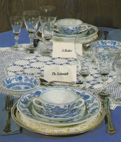 Ein festlich gedeckter Tisch mit Porzellangeschirr in floralem Blumenmuster, Kristallgläsern auf Stielen und handgeschriebenen Tischkarten mit den Namen „A. Boder” und „Th. Schmidt”. Die Gedeck-Arrangements liegen auf silbernen Platztellern, ergänzt durch Besteck und eine gehäkelte Tischdecke.
