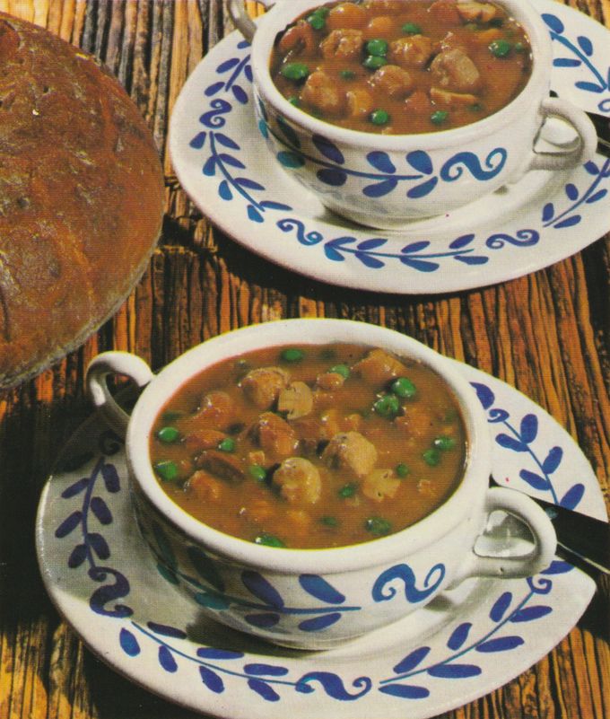 Zwei Suppentassen mit blauem Blattmuster sind gefüllt mit einer braunen Suppe, in der Champignons, grüne Erbsen und Fleischstücke schwimmen. Daneben liegt ein Laib dunkles Brot auf einer Holzoberfläche.