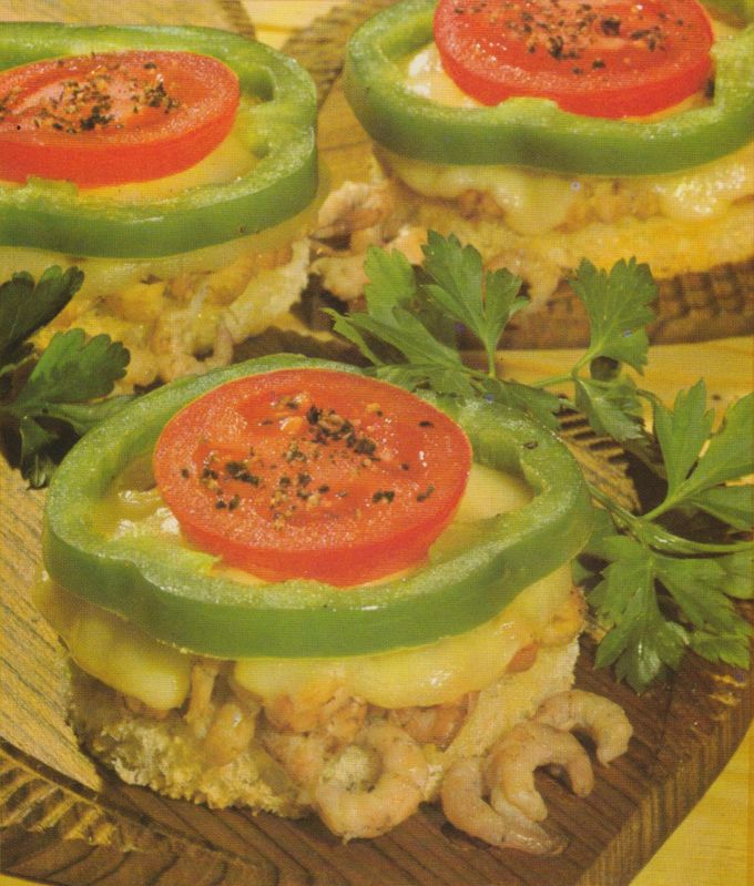 Gefüllte grüne Paprikaringe mit Tomatenscheiben und Käse auf überbackenen Toastscheiben mit Krabben, garniert mit frischer Petersilie. Die herzhaften Häppchen sind auf einem Holzbrett angerichtet und mit schwarzem Pfeffer gewürzt.