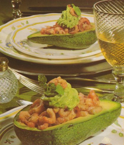 Zwei Avocadohälften gefüllt mit Nordseekrabben und garniert mit grünem Blattsalat, serviert auf Tellern mit gelbem Dekorrand. Dazu stehen ein geschliffenes Glas mit hellem Getränk und ein Salzstreuer auf dem gedeckten Tisch.