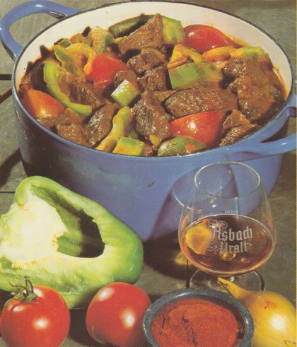 Blauer Schmortopf mit Reh- oder Hirschgulasch, dunklen Fleischstücken in brauner Sauce mit roten und grünen Paprikastreifen sowie Tomatenstücken. Davor eine angeschnittene grüne Paprikaschote, zwei Tomaten und ein Schwenker mit Asbach Uralt.