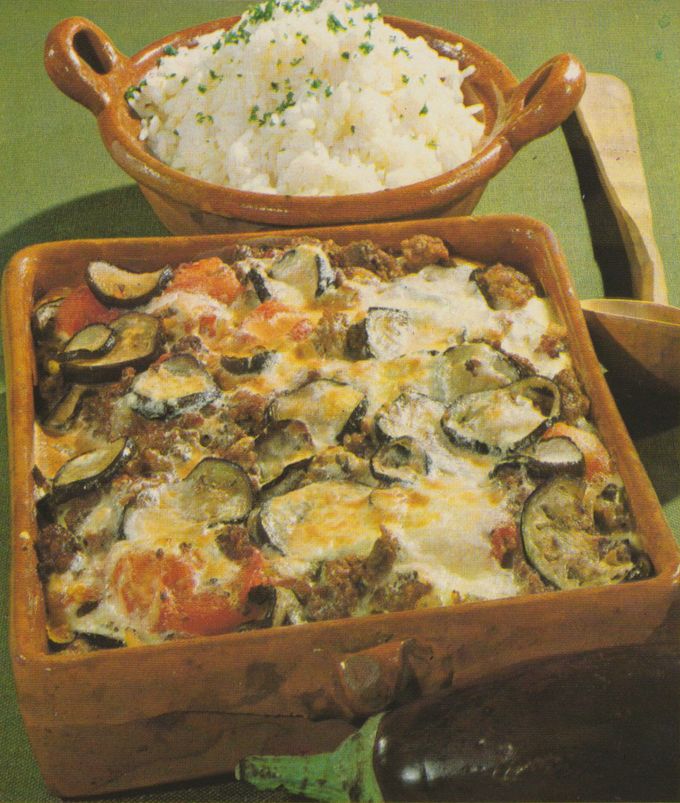 Ein Auflauf mit geschichteten Zucchinischeiben, Hackfleisch und geschmolzenem Käse in einer braunen Keramikform. Dahinter steht eine Schüssel mit Reis, garniert mit frischen Kräutern, auf einem olivgrünen Untergrund.